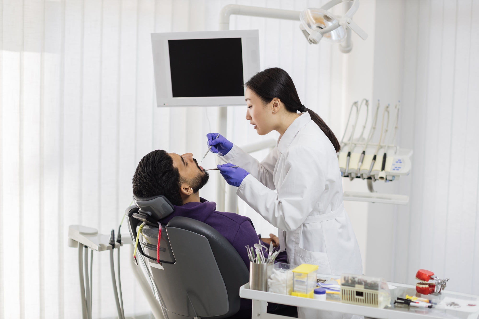 Asian dentist examines the oral cavity of young bearded handsome man